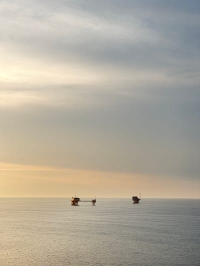 Two offshore oil rigs are visible in the calm, rippling water of the sea. The sky above is overcast with diffused light, creating a soft, hazy atmosphere. The rig on the left appears larger and more complex, with multiple structures connected by walkways. The rig on the right is smaller and more distant, consisting of a single platform. The scene is serene and industrial, with the vastness of the ocean and sky dominating the composition. The location is not specified as being in any Italian city.