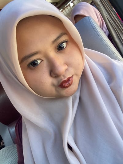 A young woman, appearing to be of Southeast Asian descent, is captured in a close-up selfie taken from a slightly elevated perspective inside a vehicle. She wears a light, possibly off-white or pale pink, hijab that frames her face and covers her head and neck. Her eyes are prominent, with visible reflections of light, and her eyelashes appear enhanced. Her dark eyebrows are well-defined. Her lips are distinctly pursed into a pout, adorned with glossy, dark reddish-brown lipstick. Her skin tone is fair, with a few faint freckles visible on her right cheek.

She is seated in what appears to be a grey, textured synthetic or leather car seat, which is visible behind her upper right side and partially behind her left shoulder. In the background, behind the grey seat, another individual wearing a pink head covering is partially visible, obscured by the seat and the main subject. Further structural elements of the vehicle's interior, including dark vertical components and light-colored pleated fabric (possibly curtains), are discernible. The scene is illuminated by natural light entering the vehicle. The context indicates this scene takes place in the city of Batu, Indonesia.
