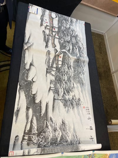 This is a painting of a snowy landscape. The scene depicts a village with several houses covered in snow. There are trees laden with snow surrounding the village. A small river or stream flows through the landscape, with snow-covered rocks and banks. A person, possibly a man walking a dog, is visible near the village.
The painting appears to be a traditional East Asian ink wash painting. The time of day is ambiguous, but the heavy snowfall suggests a winter scene. The mood is serene and tranquil, characteristic of a quiet winter day. The setting is a rural village in a mountainous or forested area, likely in China given the location context.
Visible text includes Chinese characters written in calligraphy at the top right of the painting. One inscription reads "祥雪" (xiáng xuě), meaning "auspicious snow." Another inscription to the left, in smaller characters, appears to be a poem or dedication, accompanied by a red seal. A label with text in English and possibly other languages is attached to the lower left corner of the painting.