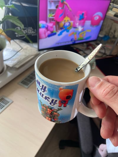 A person's hand holds a white mug filled with coffee, a spoon resting inside. The mug is adorned with a cartoon character, possibly an orange bear, and images of the Teenage Mutant Ninja Turtles. In the background, a television screen displays a colorful cartoon scene. The mug sits on a light-colored wooden surface, with other objects, including a plant and a stack of papers, visible in the background. The scene appears to be indoors, likely in a home or office setting, and does not provide any specific information about the location in Beloretsk, Russia.