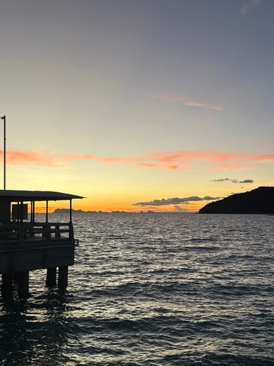 A pier extends into the ocean under a sky transitioning from deep blue to a soft orange and pink at sunset. The pier has a covered structure with visible support beams and railings. The water is choppy, reflecting the warm hues of the sky. In the distance, a silhouetted island or coastline is visible. The scene is serene and appears to be taken from a vantage point on or near the pier, looking out towards the horizon. The location is in Tong Yang, Thailand.