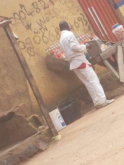 The image shows a man wearing a white shirt and white pants, with a red garment visible at his waist, standing next to a table laden with bottled drinks. The table appears to be a makeshift stand, constructed from wood. The man is looking down at the drinks, and his right hand is reaching towards them. Behind the man and the table, a rough, textured wall is visible, covered in graffiti. Some of the graffiti includes numbers and what appears to be the letters "MTN." To the right of the table, a corrugated metal structure is present. In the foreground, the ground is dusty and uneven. A white bucket with a blue lid is visible in the lower right of the image, and a smaller, dark-colored bucket is to the left of the white one. The scene suggests an outdoor market or street vendor setting in an urban environment.