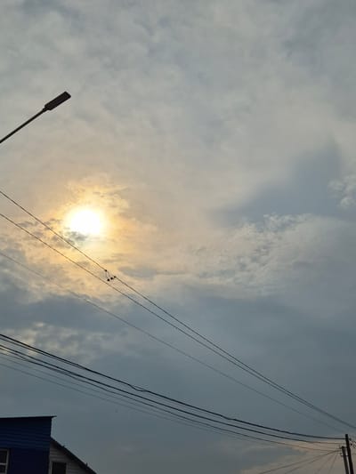 The image depicts a cloudy sky with the sun partially obscured by clouds, casting a bright glow. A street lamp extends from the left side of the frame, pointing upwards and to the right. Several power lines crisscross the sky, with a small object resembling earbuds hanging from one of the lines. In the lower left corner, the roof of a blue and white building is visible, suggesting an urban or suburban setting. The scene appears to be captured in Новоишимское, Kazakhstan. There are no people or explicit actions depicted.