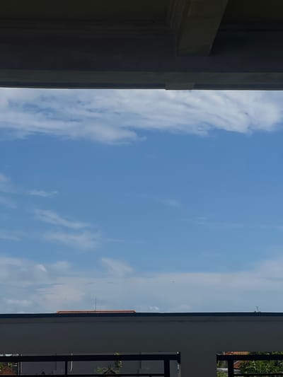 The image shows a view of the sky with scattered clouds and a blue hue, framed by architectural structures. The left side of the frame features a portion of a building with a beige color. On the right, there is a building with a white wall and a black iron fence, with hints of greenery visible through the fence. In the sky, a series of tiny dots may represent birds flying in formation. The location could be Canggu, Indonesia, but there are no specific local details to confirm. The scene is static and depicts an outdoor environment.