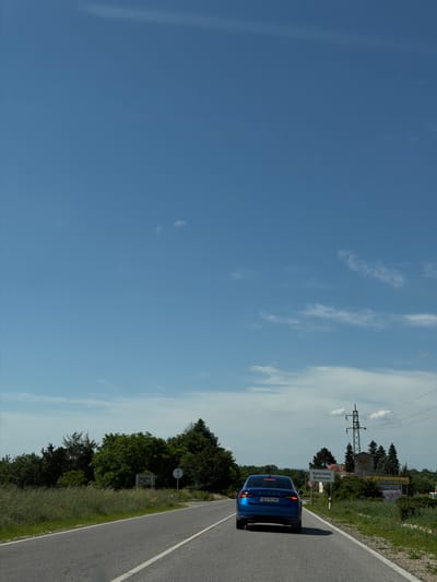 The image, rotated 90 degrees clockwise from its natural orientation, depicts a blue Skoda car on a rural road under a bright, clear blue sky with scattered white clouds. The car, bearing the license plate "CB 5191 MO" and the "SKODA" emblem on its rear, is positioned on the right side of the asphalt road, which is marked by a continuous white line.
Adjacent to the road, on the right, is a vibrant green verge with grass, bushes, and trees. A prominent white road sign stands here, clearly indicating "Брестовец" (Brestovets) in Cyrillic and "Brestovec" in Latin script, identifying the location as Brestovets, Bulgaria. Further along the road, a metal lattice utility pole with overhead power lines is visible. In the background, partially obscured by foliage, a large billboard can be seen, though its specific content is not discernible. The scene captures a sunny daytime with good visibility, suggesting fair weather conditions.