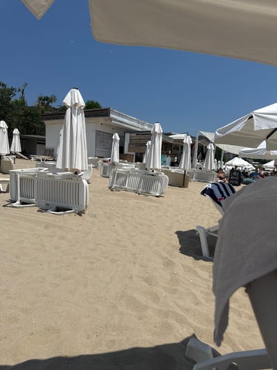 The image depicts a beach scene in Sveti Vlas, Bulgaria, on a sunny day. The foreground is dominated by sand with several white plastic beach barriers stacked and some deployed. White beach umbrellas are a prominent feature, many of them closed and covered. In the middle ground, a white building with a slatted roof and open sides serves as a beachside establishment, possibly a bar or restaurant. Several more closed umbrellas are visible around this building. Further back, more white umbrellas are open, with some beachgoers seated on loungers or beanbags. One open umbrella displays a sign that reads "PIZZA". The sky is clear and blue. The overall impression is one of a resort beach setting, with amenities prepared for visitors.