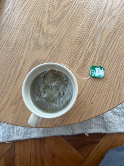 A top-down shot shows a cup of green tea steeping on a light-colored wooden table. A Clipper brand tea bag tag is visible beside the cup. A shag rug sits on one edge of the table, and hardwood flooring is visible beyond the rug. The location is not discernible from the provided image.