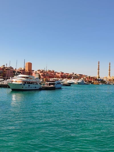 The image shows a marina in Hurghada, Egypt, on a clear, sunny day. The water is a vibrant turquoise color, with gentle ripples across its surface. Several yachts and boats are docked in the marina. In the background, a cluster of buildings with colorful facades, in shades of pink, orange, and red, line the waterfront. A tall, cylindrical orange tower stands prominently among these buildings. To the right side of the image, two tall, slender minarets of a mosque rise above the other structures, with a domed building visible behind them. The sky above is a clear, pale blue. There are no people visible in the image.