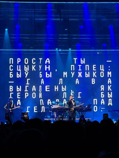 A wide shot captures a concert stage at night, bathed in vibrant blue light. A large LED screen displays bold, white Cyrillic text arranged in a grid pattern. Below the screen, performers are visible on stage. On the left, a guitarist in a dark outfit with a yellow strap plays a white electric guitar. In the center, a vocalist in a suit sings into a microphone while standing near a keyboard setup. The stage is equipped with instruments and lighting rigs. In the foreground, the silhouettes of a crowd are visible, many holding up smartphones to capture the performance. The overall atmosphere is energetic and focused on the musical act. The location is identified as Palilula Urban Municipality, Serbia.
