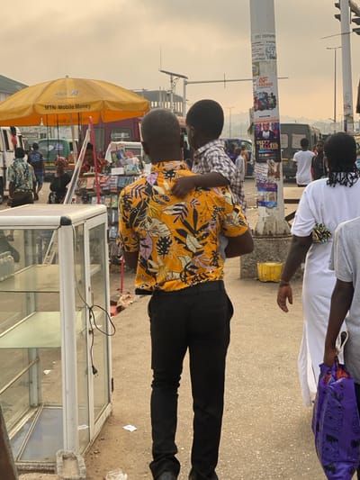 The image depicts a bustling street or market scene in Ablekuma, Ghana, under an overcast or hazy sky. In the foreground, a man, viewed from behind, wears a short-sleeved yellow shirt with black and white floral patterns and black trousers. He carries a young child on his left hip/shoulder. The child, also viewed from behind, wears a light-colored plaid shirt with their left arm resting on the man's shoulder. To the man's immediate right, a woman in a white long garment walks forward, her dark hair visible at the back, adorned with a patterned fabric accessory. Partially visible to her right is another individual holding a purple patterned bag.

To the left of the central figures, an empty, tall, white-framed glass display cabinet with two shelves stands on a rough concrete surface; an electrical cord with a two-pin plug hangs from its right side. Further left and behind, a yellow market umbrella, emblazoned with "MTN Mobile Money," shades a vendor's stall displaying numerous items, likely phone accessories, on racks. Several vehicles, including white vans and a light-colored utility vehicle, are visible in the background, suggesting traffic or parked vehicles.

To the right of the central figures, a tall, white utility pole is covered with various paper advertisements and posters, one partially legible as "CHUZ FREEMAN" and another featuring a person's face with the text "UNLY MOTED." In the far background, multiple buildings, some appearing to be under construction with exposed rebar, line the distant horizon. A solar panel is mounted on a structure overhead to the left. The ground is a light-colored, uneven paved or dirt surface with scattered debris, including small papers and a yellow object resembling a bucket. The overall scene indicates active pedestrian movement and commercial activity.