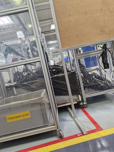 The image shows a dimly lit, industrial setting, possibly a manufacturing facility, with visible overhead lighting and a corrugated ceiling. In the foreground, there are several large, clear plastic enclosures on metal frames with wheels, appearing to be part of a production line. These enclosures contain bundles of black electrical cables and wires. One enclosure has a grey plastic bin with a yellow label that reads "Предпазна кутия" (Precautionary box) in Cyrillic script. The floor is a smooth, grey surface marked with red and yellow lines. In the background, more industrial equipment and structures are visible, including metal beams and electrical conduits. The overall impression is one of a functional, but somewhat cluttered, workspace. There are no people or animals visible in the image. It is not possible to determine the specific location within Bahovitsa, Bulgaria from the image alone.