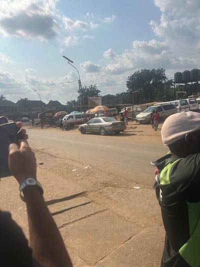 The image depicts a street scene in Igbo Ukwu, Nigeria, observed on a bright, partly cloudy day. In the immediate foreground on the left, a person's arms are visible, holding a dark-colored smartphone vertically, pointed towards the street. A white wristband is present on the left wrist. On the right foreground, the back of a person wearing a light-colored cap and a black shirt with lime green accents is partially visible, facing away from the camera.

The mid-ground features a paved road, appearing dusty, with unpaved, reddish-brown dirt shoulders on both sides. A light-colored sedan, possibly silver or beige, is positioned on the right side of the road, facing away from the viewer. Further down the road, a white van with a red stripe is visible on the left, situated under an orange market umbrella. Additional vehicles, including another white van and a silver minivan, are parked or moving further into the scene on the right.

Multiple individuals are present along the road shoulders, particularly on the right, some standing and others walking. Several market-style umbrellas in shades of orange, red, and white are spread across the scene, providing shade. A tall streetlight pole with a solar panel and an LED fixture stands prominently on the left side of the road; another similar pole is visible in the background.

The background consists of various buildings, including what appear to be single-story market stalls and multi-story structures. Green trees and dense foliage are present behind the buildings. On the far right, a structure supporting multiple dark-colored water tanks is visible against the sky. The sky above is a clear blue with scattered white cumulus clouds.