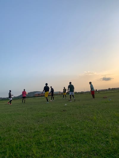The image shows a group of young men on a grassy field, likely a football pitch, in Gwafan, Nigeria. The sky is clear with a gentle gradient from pale blue to a warm, peachy hue near the horizon, suggesting it is either early morning or late afternoon. The sunlight casts long shadows, indicating a low sun angle.
There are approximately seven individuals visible, dressed in athletic wear. Most are wearing t-shirts and shorts, with some in football jerseys and shorts. Several of them are engaged in what appears to be football training or a practice session. One person in the foreground is partially visible, wearing a black t-shirt and white shorts. Further up the field, another person in a red t-shirt and black shorts is also standing. Above them, two individuals are seen in black jerseys and yellow and black shorts respectively, with one appearing to be kicking a football. Higher still, a person in a green jersey and white shorts is visible, and another in a teal t-shirt and red shorts is performing a plank. The ground is covered in well-maintained green grass, with some sparse vegetation and distant buildings or structures visible on the horizon. The overall atmosphere appears to be one of focused activity and athletic pursuit in an open, outdoor setting.