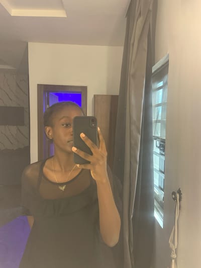 A woman in a black top is taking a selfie with a black smartphone. The selfie is taken indoors. Behind the woman is a gray curtain, a ceiling window showing a metal roof structure, and a wall with a framed window emitting blue light. The environment suggests a modern, well-lit room. The presence of these elements, although not directly indicative of Ota, Nigeria, does not exclude such a possibility. There is no specific information to pinpoint the location to Ota, Nigeria.