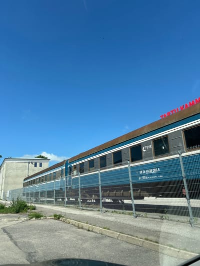This media file captures a bright, sunny day with a clear blue sky, suggesting it was taken during daylight hours. The scene features a train with dark windows and a blue stripe along its side, parked next to a wire fence. Red lettering is visible above the train, spelling out "TARTU KAM..." followed by an obscured word. On the side of the train, white text reads "EVR" and a series of numbers and symbols, including "58 26 49-38 007-5".
Adjacent to the train and fence is a paved area, likely a sidewalk or road, with some patches of grass growing along the edge. In the background, a building with a light-colored facade and dark windows is partially visible, with some greenery peeking out from its roofline. The overall setting appears to be an outdoor area, possibly a train station or maintenance yard in Tartu, Estonia, given the "TARTU" lettering. There are no people visible in the image, and the focus is on the train and its surroundings. The image is oriented such that the sky is on the left and the ground is on the right, indicating the photo was taken with the camera rotated.