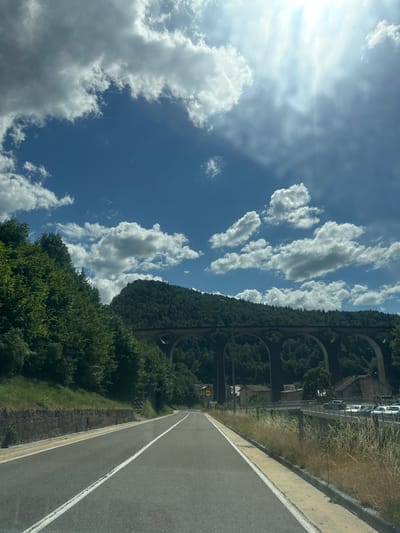 This media file depicts a scenic view of a road winding through a lush, green landscape under a bright blue sky dotted with fluffy white clouds. The road appears to be a highway with clear white lane markings. To the left of the road, a steep, tree-covered hillside rises, featuring a series of arched structures that suggest a viaduct or old aqueduct, partially obscured by foliage.
Along the upper portion of the hillside, several cars are visible parked on what might be a road or viewing area. A yellow circular sign with black text is also visible, though the text is illegible. The time of day appears to be daytime, given the bright sunlight casting shadows and illuminating the scene. The weather seems pleasant, with a mix of sun and clouds.
The overall impression is one of natural beauty and potentially historical architecture integrated into the landscape. The perspective suggests the photo was taken from a moving vehicle on the road. There is no discernible activity or event taking place, other than travel. The location is identified as Hauts de Bienne, France.