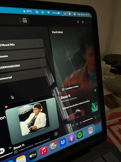 The image shows a tablet displaying a music streaming application. The screen displays the song "Beat It" by Michael Jackson, with album art featuring a picture of the artist. The user interface indicates related music videos. The tablet is placed on a desk in what appears to be a home environment. Visible on the desk are other electronic devices and household items, including a power adapter and a cup. The ambient lighting suggests an indoor setting. The date and time displayed on the tablet's status bar are March 9th, 10:15. The weather icon indicates a temperature of 25°C. The overall scene depicts a casual use of technology for entertainment within a domestic space. No specific landmarks or indications of Urbanización Encinares De Alcolea, Spain are visible in the image.