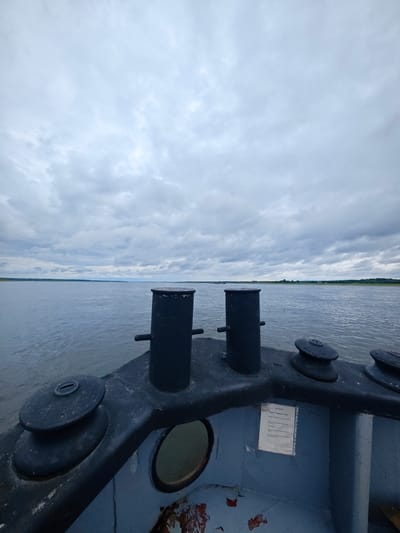 The image depicts a forward view from a boat on a large, calm body of water, likely a river, under an overcast sky. The foreground of the image is dominated by the deck of the boat, featuring two dark, cylindrical bollards and two boat winches. The deck appears weathered, with peeling paint revealing rust and primer in places. A circular porthole is visible on the side of the boat's structure. In the middle ground, the expanse of the water stretches to the horizon, where a low, tree-lined shore is visible under the cloudy sky. The overall impression is one of a quiet, possibly industrial, maritime setting. Given the context of Zotino, Russia, this scene likely represents a view from a vessel navigating the Yenisei River, a major waterway in Siberia.