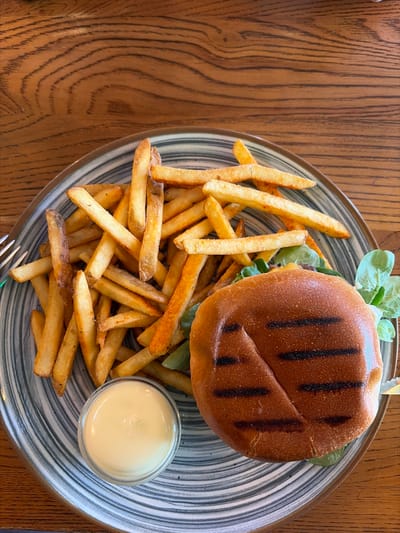 An overhead shot displays a classic fast-casual meal set on a light to medium-brown wooden table with distinct grain patterns. The central subject is a circular plate featuring a burger, a generous portion of french fries, and a small container of sauce.
The burger, positioned towards the top right of the plate, has a toasted bun with prominent dark grill marks, suggesting it was grilled. Visible beneath the bun are fresh green lettuce leaves and a hint of the burger patty. Surrounding the burger is a large serving of golden-brown french fries, varying in length and thickness, some appearing crisp.
In the bottom right corner of the plate, a small, clear glass or plastic cup holds a creamy, pale yellow sauce, likely mayonnaise or aioli, intended for the fries. The plate itself is circular with a notable design: a wide rim featuring concentric, wavy stripes in shades of blue-gray and white, contrasting with a lighter, solid-colored center.
The overall scene suggests a prepared meal, possibly lunch or dinner, ready for consumption in a restaurant or cafe setting in Essen, Germany. The lighting is bright and even, indicating an indoor environment, potentially illuminated by daylight or good artificial lighting. A small part of a metallic fork is visible on the bottom left edge of the plate. No people are visible, and no text is present in the image.