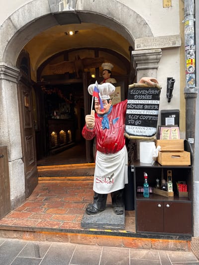 The image shows the exterior of a restaurant, likely in Ljubljana, Slovenia, given the context clues. A life-size statue of a chef stands prominently outside the entrance. The chef is dressed in a red jacket, white apron, and chef's hat, and is holding a Slovenian flag and giving a thumbs-up gesture.
In front of the statue is a display board listing "SLOVENIAN DISHES," "HOUSE BEER," "DOMAČE PIVO," "ILLY COFFEE TO GO," and "ILLY KAVA ZA S SEBOJ." To the right of the display board is a shelf unit containing various items. These include two wooden crates, a roll of paper towels, cleaning spray, bottles of olive oil, and packets of condiments.
The restaurant entrance is an arched doorway with wooden doors, and the interior is dimly lit, with glowing lights highlighting a staircase leading inside. Above the entrance, a stone archway frames the scene. A sign on the wall above the statue reads "SELFIE SPOT" with the hashtag "#gostilnasokol," indicating the restaurant's name is Gostilna Sokol. The overall setting suggests a welcoming and traditional dining establishment.