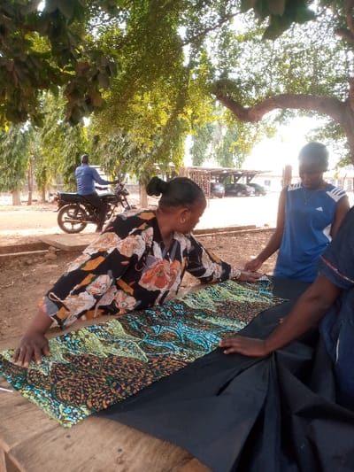 An outdoor scene in Agban, Nigeria, features three individuals interacting with fabric under large trees, with a male on a motorcycle in the background. In the foreground, a woman with dark skin, wearing a black shirt patterned with large orange, white, and green flowers, leans over a wooden table. Her hands are placed on a vibrant green, blue, and brown patterned fabric, which lies over a solid black fabric. To her right, a second woman, also with dark skin, wearing a blue sleeveless Adidas top with white shoulder stripes, stands and looks down at the fabric, her hands positioned on it. A third person's right arm and hand are visible on the far right, resting on the black fabric. In the mid-ground left, an adult male in a blue long-sleeved shirt is seated on a stationary black motorcycle, facing away from the viewer towards the right. The background includes more slender trees, a dirt path, and a metal-roofed open shelter covering several parked dark cars, indicating a developed area within the city. The scene is illuminated by natural light filtering through the tree canopy.