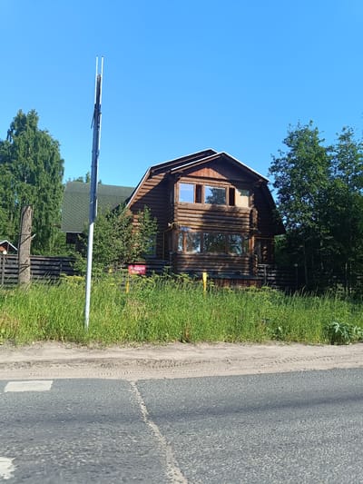 A log cabin is situated in a rural setting with a clear blue sky. The house has a dark wood exterior with several windows on different levels. A green roof is visible. The cabin is surrounded by lush green grass and trees. A wooden fence runs along the property line. A tall, thin pole stands in front of the cabin, its purpose unclear.
A red sign with white text is attached to the fence in front of the house. The text on the sign is in Cyrillic script and appears to be an advertisement or a sale notice. The time of day is likely daytime, given the bright sunlight casting shadows. The weather appears to be clear and sunny. The location is implied to be Arkhangelsk, Russia. The scene suggests a peaceful, possibly residential area, with the house looking like a home or a vacation property.