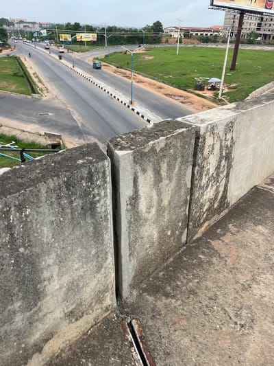 The image shows an elevated view of a section of road in Osogbo, Nigeria. The camera is positioned above a concrete barrier, with the road stretching into the distance. A median with alternating black and white stripes divides the road. Vehicles, including cars and motorcycles, are visible on the road. Buildings, billboards, and green spaces line the road. A small market area is visible to the right of the road in the middle distance. The sky appears overcast. The concrete barrier in the foreground is stained and shows signs of weathering.