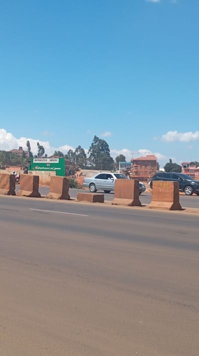 A road with concrete barriers divides the foreground, leading to a scene with a green sign reading "Gatuguta's House" and "Salaricom Mpesa." Cars are driving on the opposite side of the road: a silver car and a black SUV are prominent. Buildings and trees populate the background under a clear, blue sky with scattered clouds. The scene depicts a roadside view with local advertisements.
