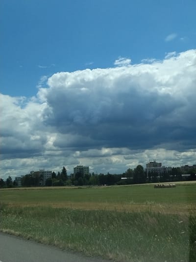 This image captures a vast expanse of sky filled with dramatic, dark storm clouds dominating the upper portion of the frame. Below the clouds, a line of apartment buildings and trees forms a distant horizon. In the foreground, a green field stretches across the scene, with a paved road visible on the left edge. Several hay bales are scattered across the field in the middle ground. The overall impression is a partially overcast day with a hint of impending weather over a semi-urban landscape. The location is the city of Tábor, Czechia.