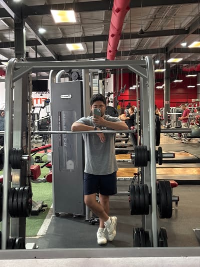 An upright mirror selfie captures a man in a gym setting. The man, wearing a gray t-shirt, dark shorts, and white sneakers, is reflected holding a smartphone to take the picture. He stands in front of a large, gray weight machine, possibly a Smith machine or a selectorized machine, where the reversed text "NAUTILUS" and "DUAL GLUTE / CALF" is visible.
Around the man, numerous black weight plates are stacked on integrated racks, and additional gym equipment is seen in the background. Other gym patrons are visible in the reflection, engaged in their workouts. One person in a red top is near a machine, and another with red hair is near a weight bench. The gym features red accent walls, a high ceiling with exposed metal beams, ductwork, and rectangular overhead lights. A large, red padded ventilation duct runs horizontally across the upper left. The flooring includes sections of green artificial turf with a white line. Partially visible reversed text on a banner in the background includes "LIFT", "ZODIA", and "POWER". The overall impression is an active fitness facility in Cholula, Mexico.