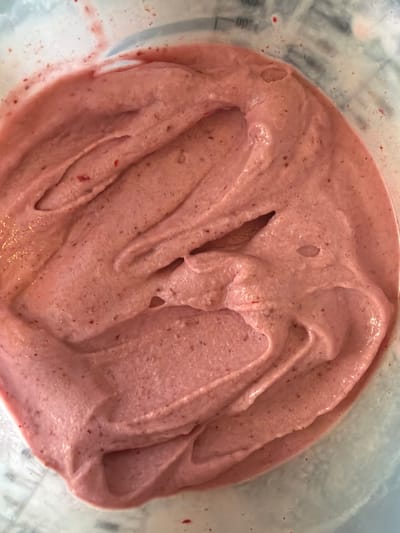 This media file is a close-up, overhead shot of strawberry ice cream or a similar frozen dessert. The dessert is pinkish-red in color and has a soft, swirled texture, indicating it has recently been churned or scooped. Tiny red flecks, presumably pieces of strawberry, are visible throughout the dessert. The lighting is bright and even, suggesting it was taken indoors during the daytime. The background is out of focus but appears to be the inside of a clear container or bowl, possibly a blender jar, with some faint measurement markings visible on the left side. There is no discernible time of day, weather, or location cues other than the generic domestic setting. The overall impression is one of a freshly made, appealing dessert. No people or specific event are depicted.