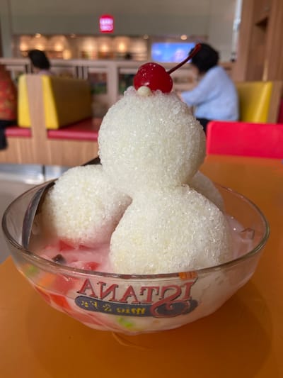 A close-up, eye-level shot features a dessert in a clear glass bowl filled with a pink liquid and colorful fruit pieces. The dessert itself consists of four scoops of white, granular ice, with a maraschino cherry and a white bead topping the central, uppermost scoop. A silver spoon is partially visible on the left side of the bowl. The bowl rests on an orange table. In the blurred background, several people are seated at tables in a restaurant setting. One person in the mid-ground wears a light blue shirt, and another person in the foreground-left is partially visible wearing a patterned orange shirt. Yellow and red seating are also visible in the background. The restaurant appears to be "TATA" based on the text visible on the bowl. The overall scene suggests a dessert being served in a casual dining establishment.