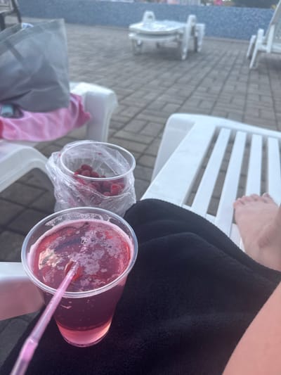 The image depicts an outdoor scene, likely a poolside area with lounge chairs, on a paved surface. In the foreground, a clear plastic cup filled with a pinkish-red beverage, possibly a cocktail or juice, is visible. A pink straw extends from the drink. Next to the cup, there is a clear plastic bag containing several red berries, likely raspberries. A black towel is draped over a white lounge chair in the foreground, partially obscuring the view of a person's feet and legs. In the background, other white lounge chairs and poolside furniture are visible, along with a body of water, possibly a swimming pool. The overall impression is one of relaxation and leisure. The image does not provide any specific visual cues that would allow for the precise identification of the location within Makiivka, Ukraine.