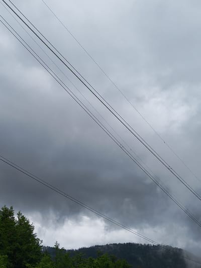 The image depicts a cloudy sky with electrical power lines running diagonally across the frame. Dense green trees are visible on the right side of the image, suggesting a wooded or mountainous area. The general overcast condition creates a muted color palette, emphasizing shades of gray and green. The image appears to have been taken in Barziya, Bulgaria.