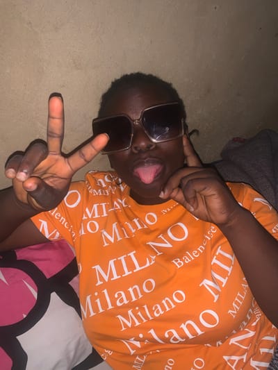 A young Black woman is pictured indoors, wearing large, square-framed sunglasses and an orange t-shirt with a repeating white pattern. She is making a peace sign with her left hand and sticking out her tongue. Her right hand is raised to her ear. The background appears to be a neutral-colored wall. There is a pillow with a pink and black circular pattern visible on the left side of the frame, and a dark fabric, possibly a blanket, is draped over her right shoulder. The lighting suggests this is an indoor setting at night or in a dimly lit room, possibly in Agban, Nigeria.
