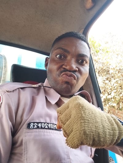 A male individual is depicted in a close-up, selfie-style photograph, situated inside a vehicle in Ejule, Nigeria. He presents with dark skin, dark hair, and a dark mustache. His eyes are wide open, and his lips are pursed forward in an exaggerated grimace. He is wearing a light pinkish-beige collared uniform shirt with visible shoulder epaulets and a dark rectangular patch on the left chest pocket, displaying white characters that appear to read "8S FOSFUPU". His right hand is raised, formed into a loose fist, and covered by a light tan, fingerless knitted glove. The vehicle's interior is visible, including a light beige fabric headliner and a dark seat headrest. Through the vehicle's windows, a light blue object is partially visible on the left, and sparse, light-colored foliage or dry vegetation is visible on the right, indicating an outdoor, sunlit environment.