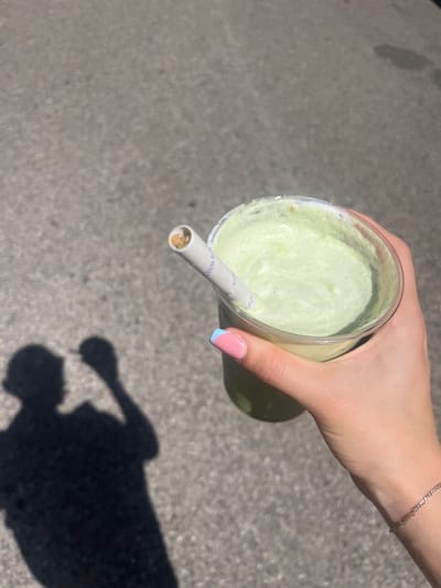 A top-down shot shows a hand holding a clear plastic cup filled with a light green beverage, likely a smoothie or blended drink. A white paper straw with brown residue inside is inserted into the cup. The hand has a pinky fingernail with a two-tone design: mostly pink with a blue accent at the tip. A delicate silver bracelet adorns the wrist. The background is a gray concrete surface. A shadow of a hand holding a cup is visible in the lower right quadrant. Based on the shadow's clarity and direction, the photograph appears to have been taken in daylight. No information suggests a location in the city of Saarbrücken, Germany.