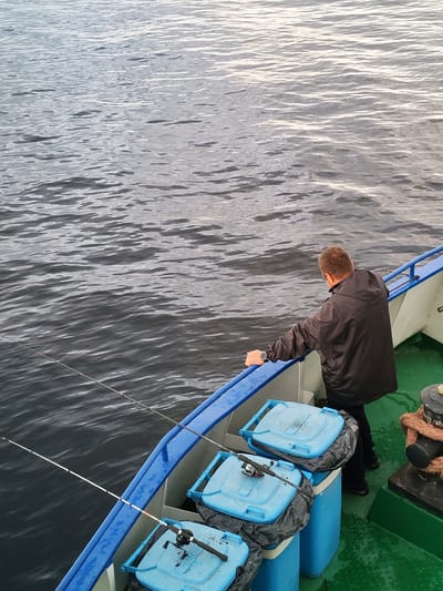 A man is fishing from the deck of a boat in Shcheleiki, Russia. The boat has a blue railing and green deck. There are three blue bins with fishing rods resting on them. The water is dark and choppy. The man is wearing a dark jacket and has a watch on his left wrist. He is leaning over the railing, looking at the water.