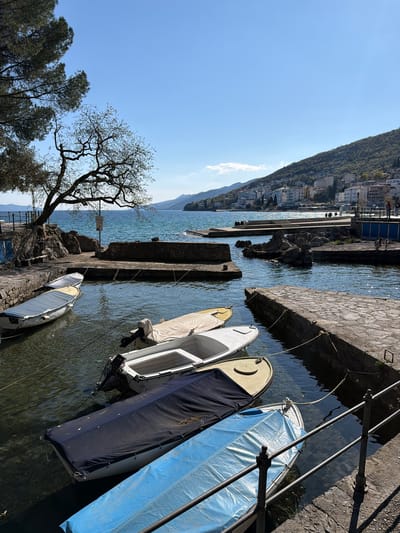 The image depicts a harbor scene in the city of Grad Opatija, Croatia, with several small boats moored alongside a stone quay. The boats are of varying sizes and colors, some covered with protective tarps. The quay is constructed of rough-hewn stone blocks. In the background, a mountain rises steeply from the water's edge, with buildings visible along the coast. A sparse tree stands near the quay. The water is calm and clear, with reflections of the sky and surrounding landscape. The sky is blue and cloudless, indicating a sunny day.