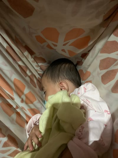 A baby sleeps peacefully in a hammock. The hammock fabric has a pattern of orange and white shapes. The baby is covered with a yellow blanket and is wearing a pink garment with writing on it. The writing on the garment is partially visible and appears to be in English. The background of the image is a close-up of the hammock's fabric, suggesting the photo was taken from within or near the hammock. The lighting suggests an indoor environment, possibly at night or in a dimly lit room. The image is a personal photograph, likely taken by a caregiver. The location in Kiblawan, Philippines, indicates a residential setting within this municipality.
