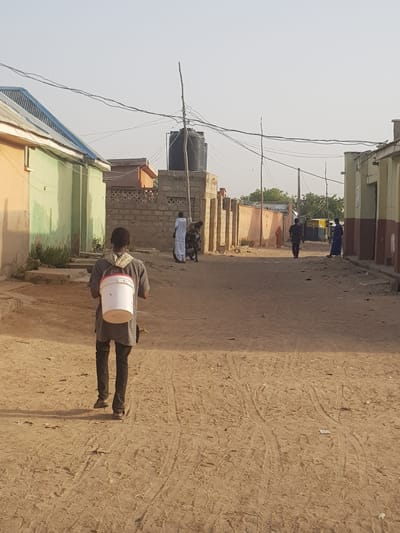 The image captures a street scene in Dabawa, Nigeria. The street is unpaved, revealing sandy, dirt ground with tire tracks visible. A person is walking away from the viewer, carrying a white bucket with red trim over their shoulder. In the background, several people are visible further down the street. Buildings line both sides of the street, constructed from a mix of concrete and brick. A large, black water tank sits atop one of the buildings. Overhead, a tangle of power lines crisscrosses the sky.