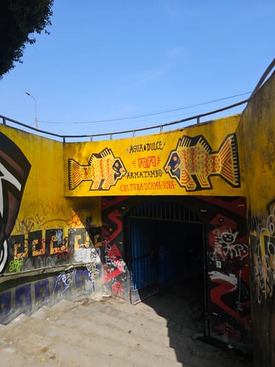 This image shows an underpass in Chorrillos, Peru, possibly a pedestrian or railway underpass. The underpass has been heavily decorated with graffiti, including murals depicting fish in a stylized, geometric design. A yellow and red color scheme is prominent. Text in the graffiti reads "AGUA DULCE," "MATAMBO," and "CULTURA ICHMA VIVA." The underpass itself appears to be made of concrete. There is a metal gate or fencing visible at the end of the underpass. The sky above is blue, and some green foliage is visible on the left side of the frame. The area around the underpass appears to be urban. The underpass's interior is dark.