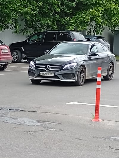 The image shows a dark-colored Mercedes-Benz C-Class sedan parked on an asphalt surface. The car is facing slightly to the right, and its front license plate is visible, displaying the text "P953KK 799". The car has its headlights on. In the background, a black SUV is parked behind the Mercedes, and further back, part of a red car is visible on the left. Trees with dense green foliage form a backdrop above the cars. A red and white striped traffic cone is positioned in the foreground to the right of the Mercedes. The scene appears to be an outdoor parking lot, possibly in a suburban or semi-urban area. The lighting suggests it is daytime, and the overcast sky indicates cloudy weather. There are no obvious emotional cues from the scene. The context provided indicates the location is Pavlovsky Posad, Russia.
