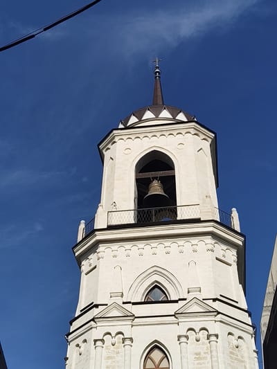 A low-angle photograph captures the upper two-thirds of a white, octagonal church bell tower against a deep blue sky with minimal, faint cloud cover. The tower culminates in a dark brown, conical spire topped with a small, metallic Orthodox cross. Below the spire, a dome-like cap features alternating white and dark brown triangular segments.

The uppermost octagonal section of the tower, functioning as a belfry, is constructed from white stone or stucco. It exhibits open, pointed arched portals, with a large, ornate brass bell visibly suspended from dark wooden beams within one of the arches on the front face. Below the belfry arches, a black metal balcony railing with a diamond mesh pattern encircles the structure. This section is adorned with small, rounded decorative elements (corbels) and stylized pilasters.

Below the belfry, another octagonal section is visible, featuring a band of small, regularly spaced semicircular decorative elements. Further down, pointed arched windows, some with visible panes, are set into the white stone, flanked by slender, vertical architectural decorations resembling miniature pinnacles or pilasters. The tower's surface shows minor signs of weathering and texture.

A thick, black cable is visible diagonally across the extreme upper left corner of the frame. A sliver of a dark, reflective surface, possibly from an adjacent building, appears on the far right edge. The lighting suggests direct sunlight, creating subtle shadows that define the architectural relief. The scene is located in Bykovo, Russia.