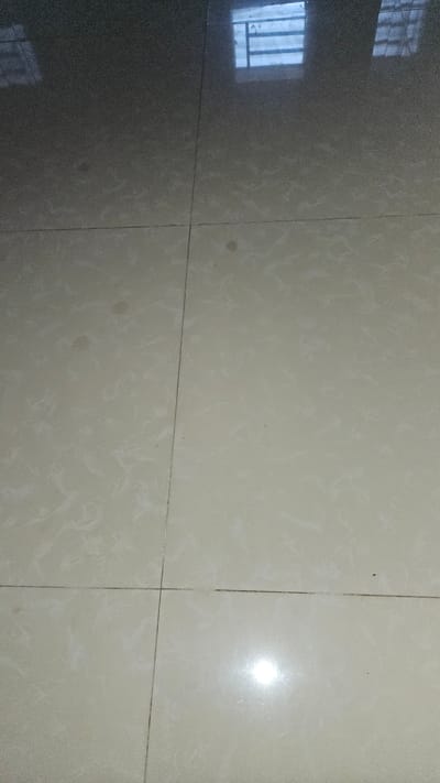 The image shows a section of tiled flooring. The tiles appear to be light-colored with a subtle mottled pattern. Grout lines are visible between the tiles. On the left side of the image, there is a dark reflection of what appears to be a window with bars. The reflection indicates the surrounding environment may be in Cam Đức, Vietnam. The flooring is clean and uncluttered. There are no people or other objects present.