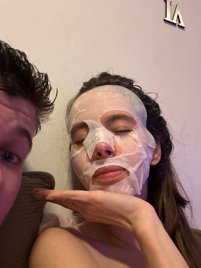 This is a low-angle, close-up image featuring two individuals, likely taken indoors during the evening due to the dim, artificial lighting which casts subtle purple/pink hues.
In the foreground, on the bottom left, is the partial face of one person. Only their left eye, eyebrow, and part of their forehead are visible. They have dark, short, spiky hair that appears somewhat damp or styled. Their eye is open and directly looking towards the camera, suggesting they might be taking the photo or are actively engaged with the person taking it.
Dominating the central and upper right portion of the image is another person, seen from a slightly lower and more distant angle. This individual has long, dark, wavy hair that also appears somewhat damp. Their face is covered by a white sheet mask, which has cutouts for the eyes, nostrils, and mouth. Their eyes are closed, indicating a state of relaxation. Their lips are slightly parted, revealing the inside of their mouth. A bare arm is visible, bent at the elbow, with a hand resting near their shoulder. A ring with a visible stone is on one of their fingers. Part of their upper torso or shoulder is also visible. This person is engaged in a skincare routine with the face mask.
The background consists primarily of a light-colored wall, possibly white or light grey. In the upper left corner of the image, attached to the wall, are two metallic, silver-colored letters that read "VI". These letters are raised and cast a slight shadow on the wall. Below the arm of the person with the mask, on the right side of the image, there is a visible section of a textured, ribbed fabric, possibly a cushion, blanket, or part of a sofa, in a dark brown or grey color. The overall atmosphere suggests a relaxed, personal, and intimate moment.