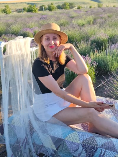 A woman with blonde hair and wearing a straw hat, black t-shirt, and white shorts sits on a bed in a lavender field. The bed has a white metal frame and is covered with a patchwork quilt and sheer white fabric draped over it. The woman is holding a smartphone and looking towards the camera with a smile. The background shows a rolling landscape with lavender fields and trees. The scene is illuminated by natural light, suggesting it is daytime. The location is specified as Nechaiane, Ukraine.