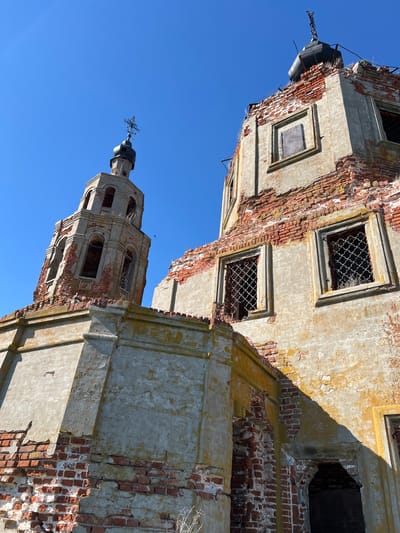 The image depicts the decaying remains of a brick and concrete structure, possibly a church, in Шуран, Russia, under a clear blue sky. The structure is heavily weathered, with crumbling brickwork and dilapidated walls. Window openings are visible, some with remnants of iron grates. A tower topped with a cross is present. The image is shot from a low angle, emphasizing the height and decay of the structure.