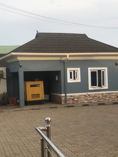 The scene depicts the exterior of a single-story building in Egan, Nigeria, under an overcast sky. The building features blue-grey walls, a dark-tiled hip roof with white fascia and gutters, and a lower wall section adorned with multi-toned stone veneer. On the right, two white-framed windows are present; the larger window's right pane is partially open outwards, while the smaller left pane remains closed. A dark drainpipe runs vertically down the building's right side. On the left, a recessed alcove contains a large yellow industrial generator branded "CAT," positioned behind a black control panel. A red fire extinguisher is mounted on the wall within this alcove. To the immediate left of the alcove, two red plastic containers rest on the paved ground. In the immediate foreground, a partial view of a silver-toned metallic railing with a spherical finial is visible. The ground is paved with interlocking tan and grey pavers. A single bird is perched on the peak of the building's roof. Two thin overhead wires traverse the upper left portion of the sky. No human figures are present in the image.