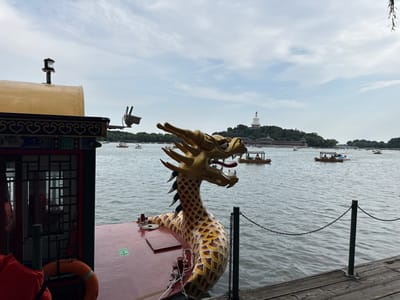 This media file depicts a scenic view of a lake with several boats and a white pagoda in the distance. The foreground features the front of a boat adorned with a prominent golden dragon head. The dragon's scales are detailed in red and yellow, and its mouth is open, revealing fangs and a red tongue. The boat also has a railing and what appears to be an orange life preserver hanging on the left side.
In the background, the lake is dotted with numerous small boats, some carrying passengers. The boats appear to be traditional Chinese vessels. Beyond the lake, a lush green hillside rises, topped by a striking white stupa, which is a prominent landmark. The sky is overcast with white clouds, suggesting a cloudy or possibly overcast day. The overall setting appears to be a park or recreational area, likely a popular tourist destination.
The image conveys a sense of tranquility and cultural richness. The presence of the dragon boat suggests a traditional or festive atmosphere. The location is Xicheng District, China, and the white stupa is likely the White Dagoba on Qionghua Island in Beihai Park. There is a green arrow symbol visible on the deck of the boat in the foreground, but no other legible text is present. The time of day is difficult to ascertain precisely due to the overcast conditions, but it appears to be daytime.